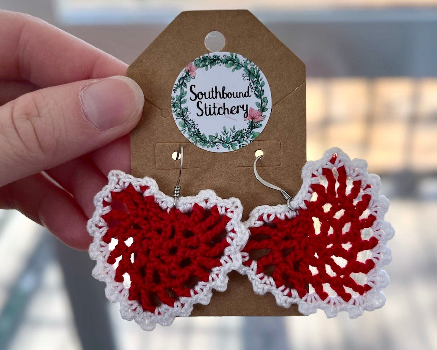Red and white crochet earrings on a Southbound Stitchery card held by a hand.
