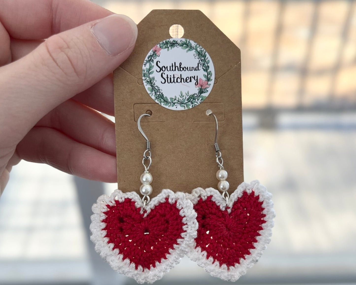 Red heart-shaped earrings on a Southbound Stitchery card held by a hand.