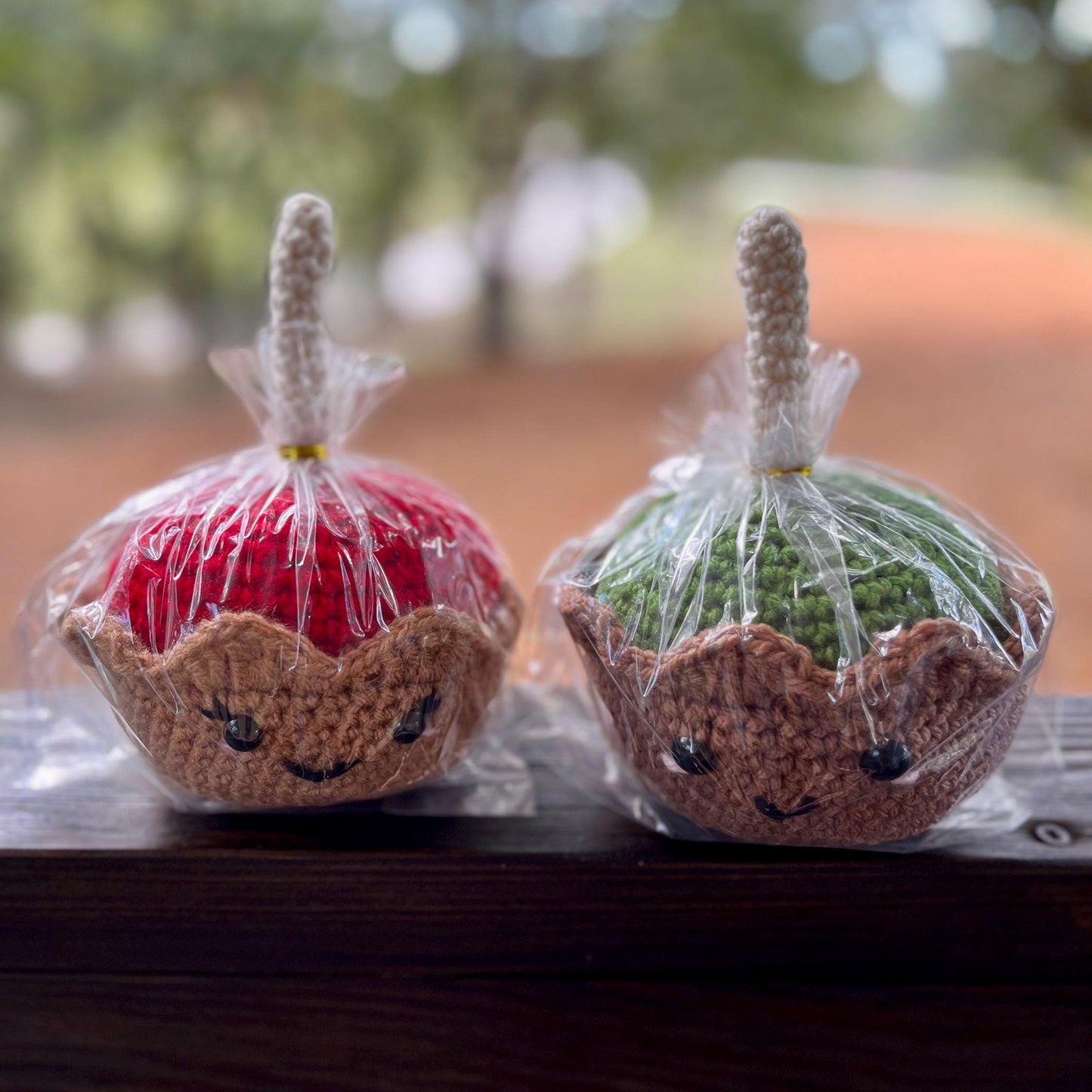 Two crochet apples in clear plastic wrap on a wooden surface with a blurred natural background.