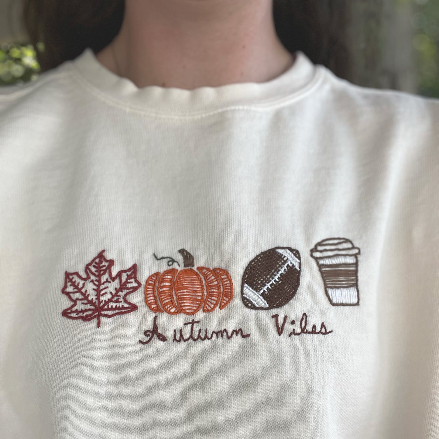 White sweatshirt with embroidered autumn-themed design featuring leaves, pumpkins, and text.