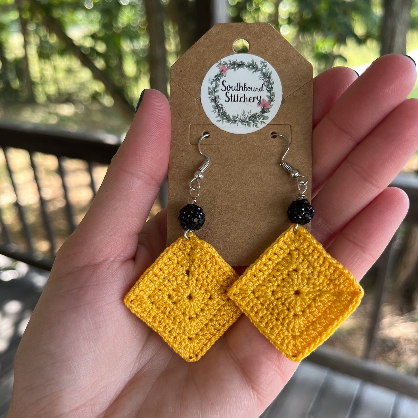 Yellow crochet earrings on a Southbound Stitchery card held in a hand with a blurred outdoor background.
