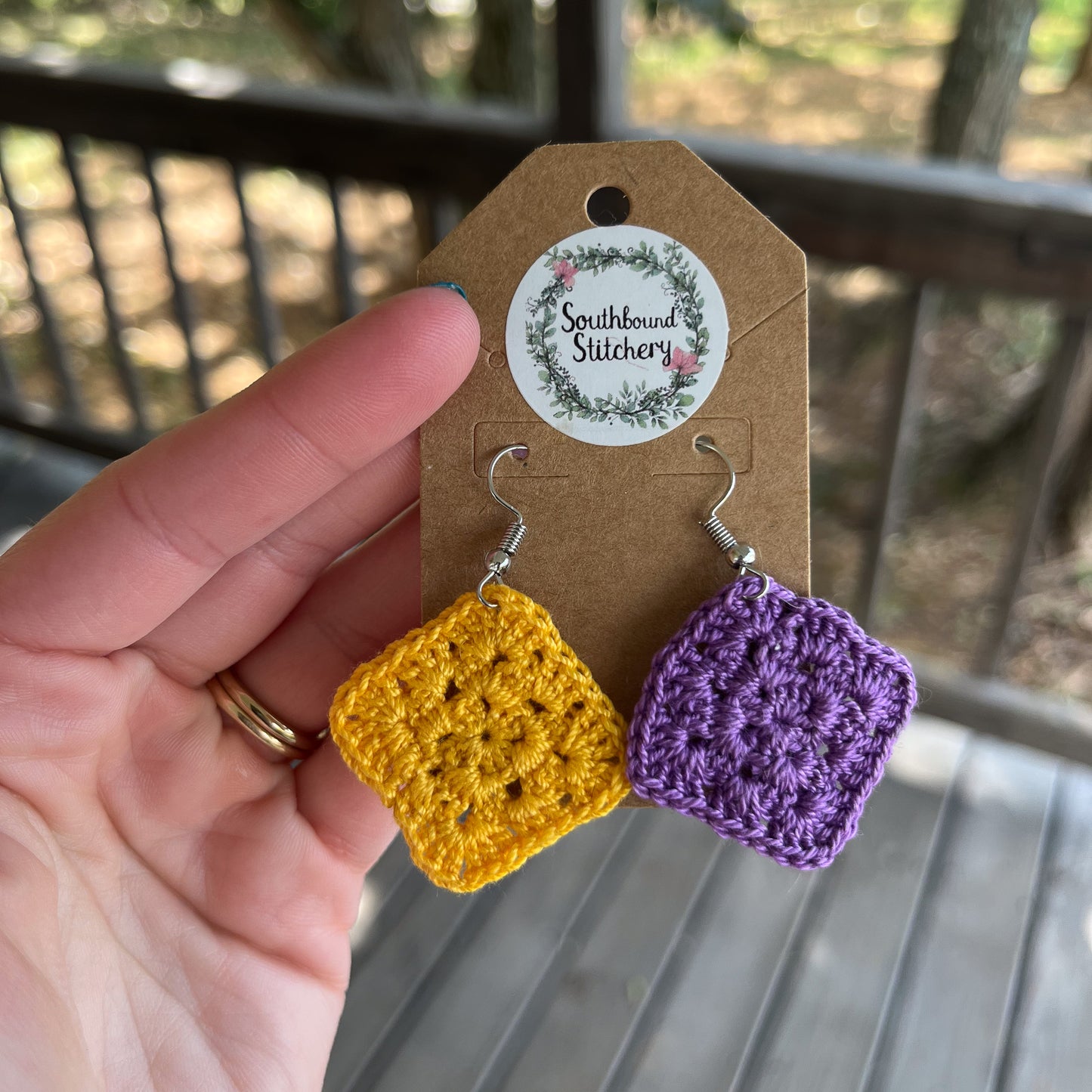 Crocheted square earrings in yellow and purple on a Southbound Stitchery card, held by a hand.