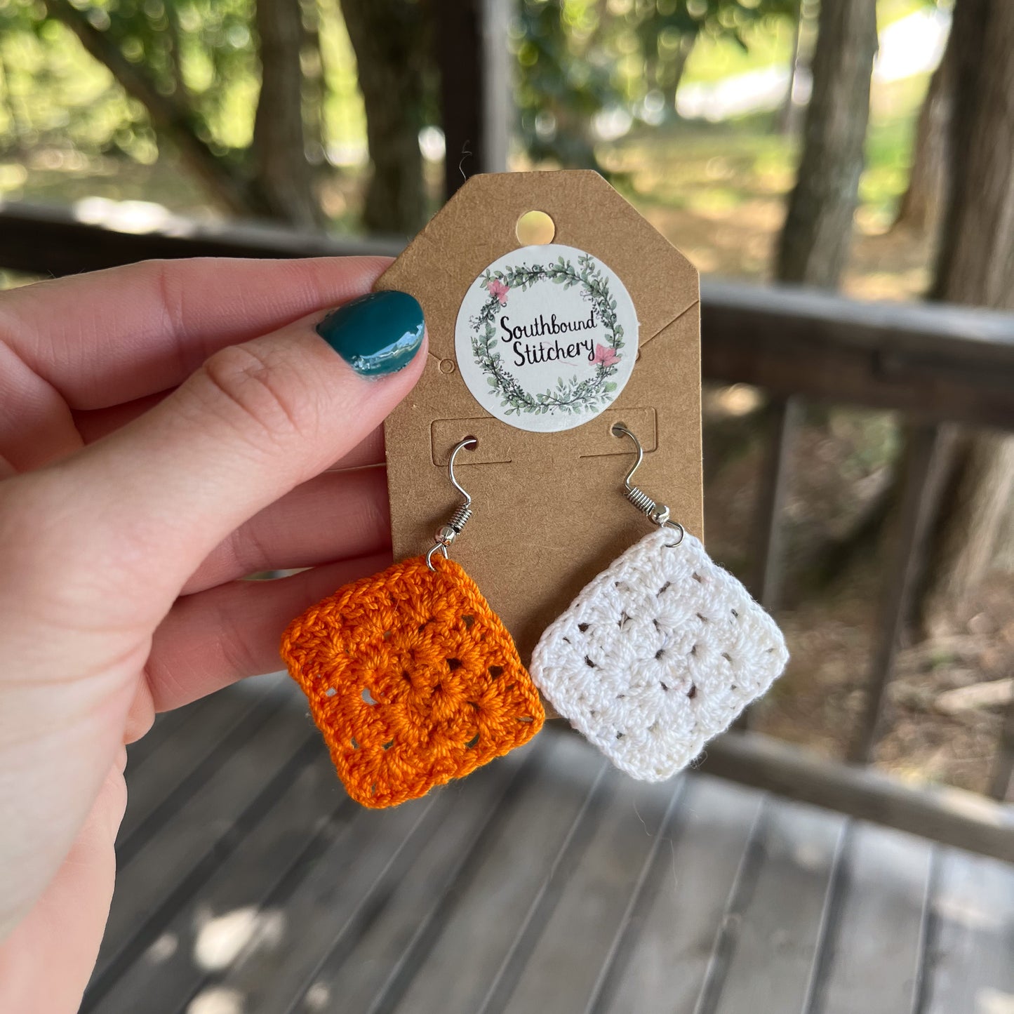 Hand holding orange and white crochet earrings on a Southbound Stitchery card with a blurred natural background.