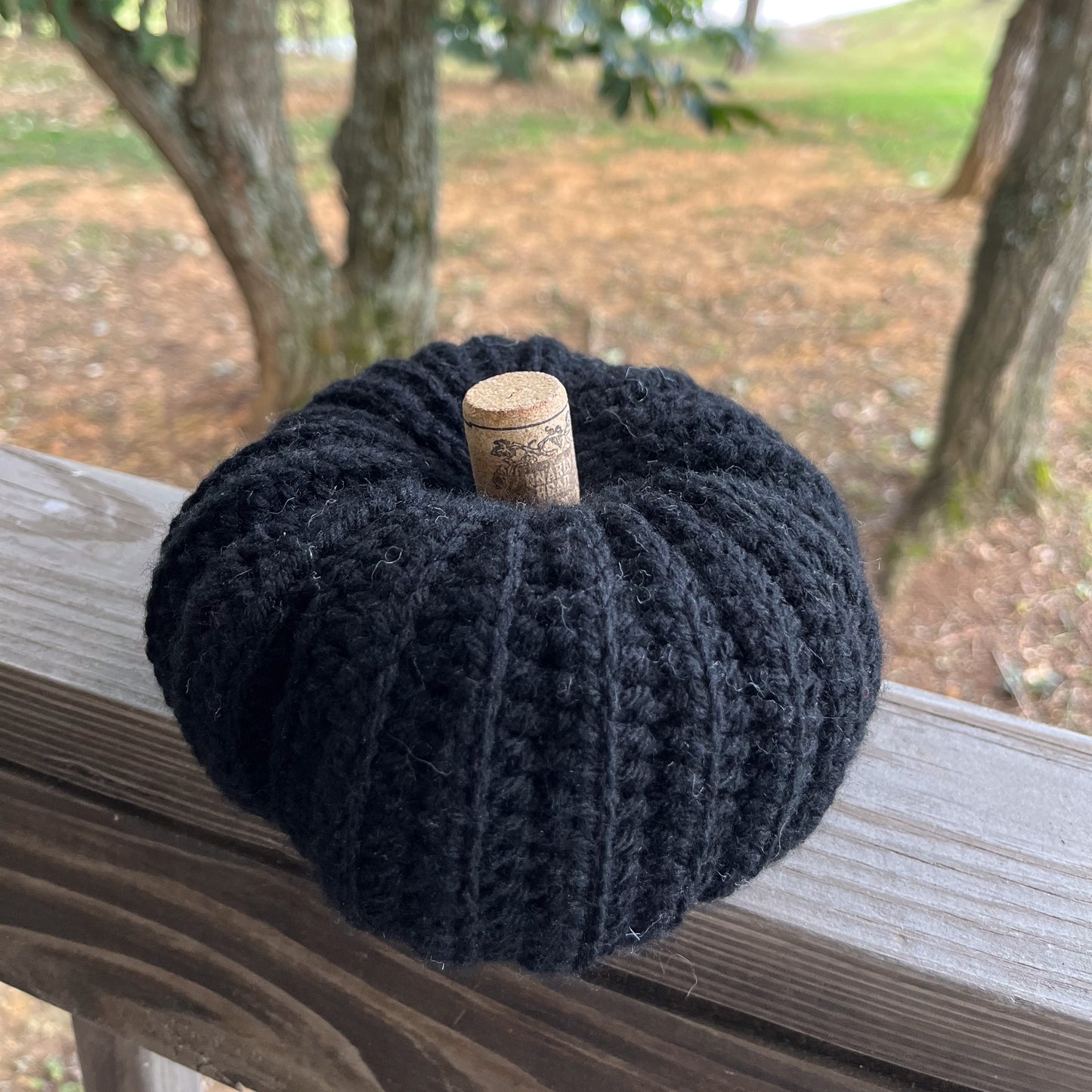 Black Crocheted pumpkin with a cork on top, placed on a wooden surface outdoors.