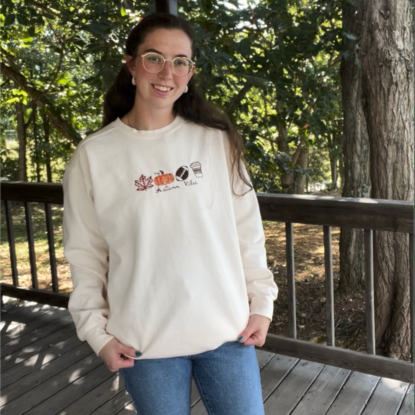 Person wearing a white sweatshirt with a design on a wooden deck with trees in the background