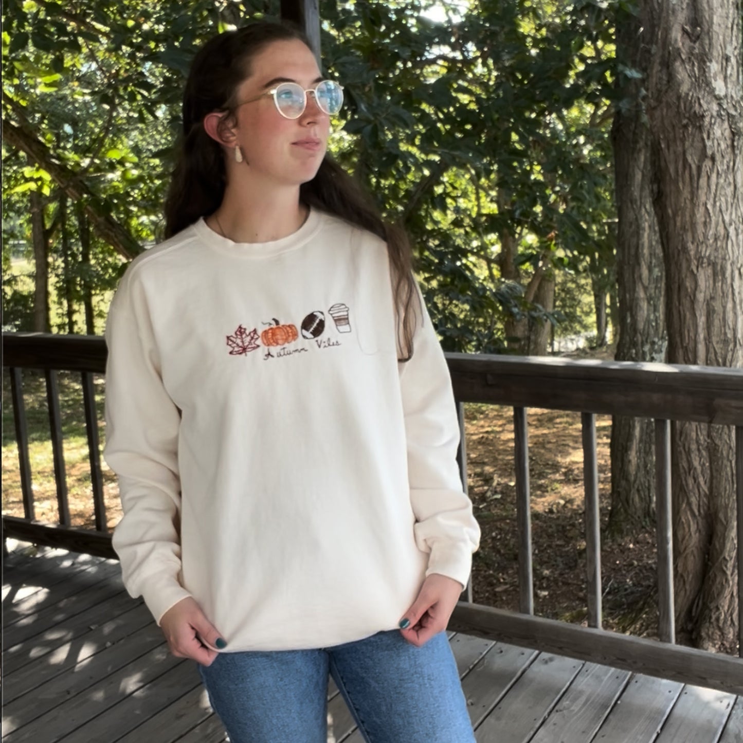 Person wearing a cream sweatshirt with graphics and text on a wooden deck with trees in the background