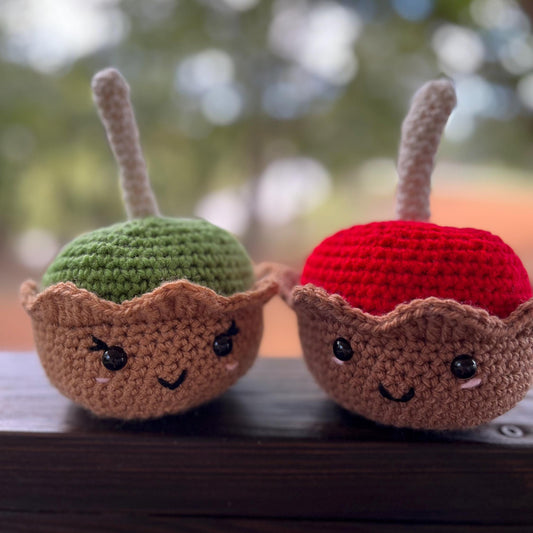 Two crochet apples with faces on a wooden surface, blurred greenery in the background