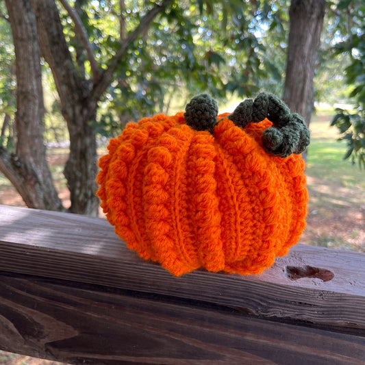 Crocheted pumpkin on a wooden surface with trees in the background